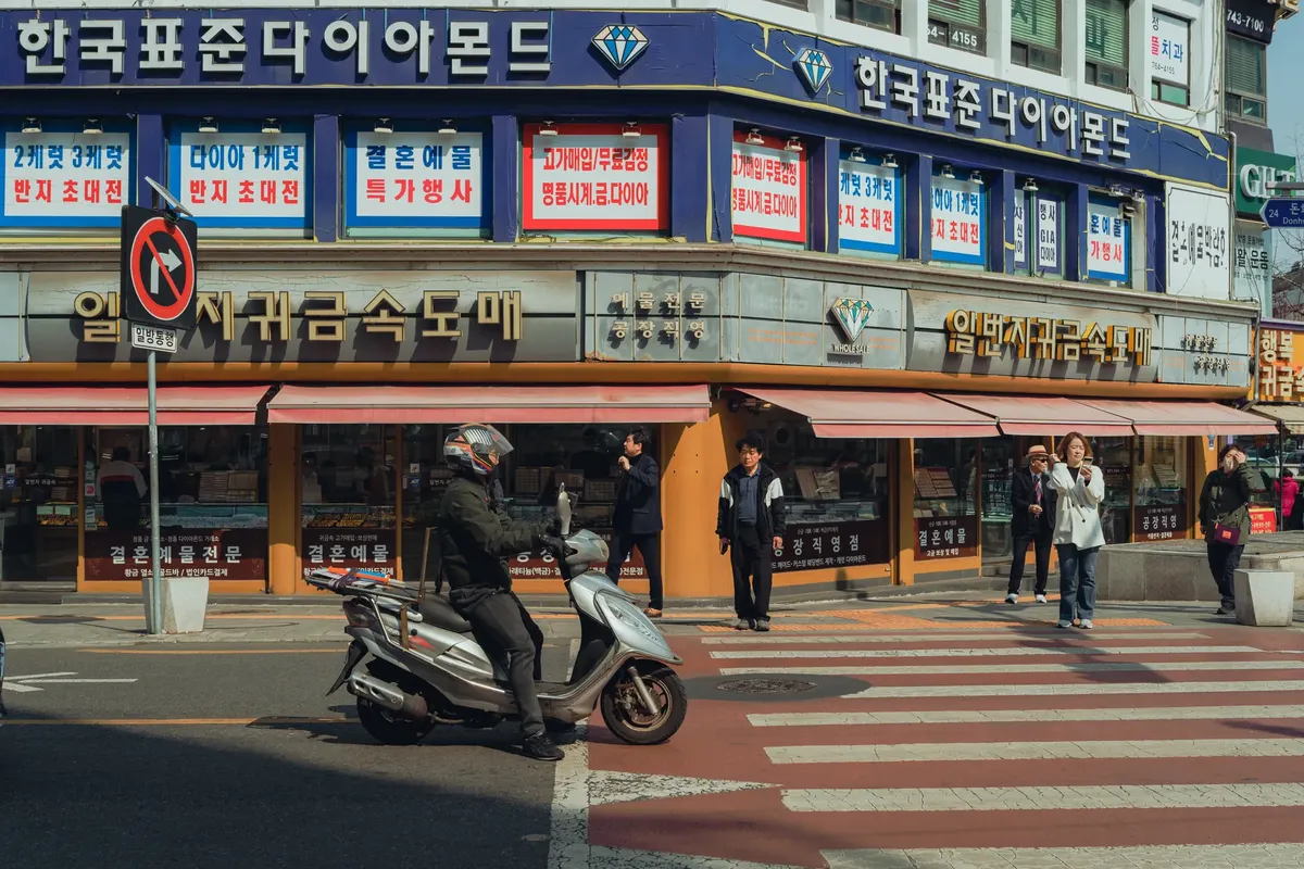 Urban street scene in Seoul featuring a motorcyclist and pedestrians with Korean shop signs in the background.